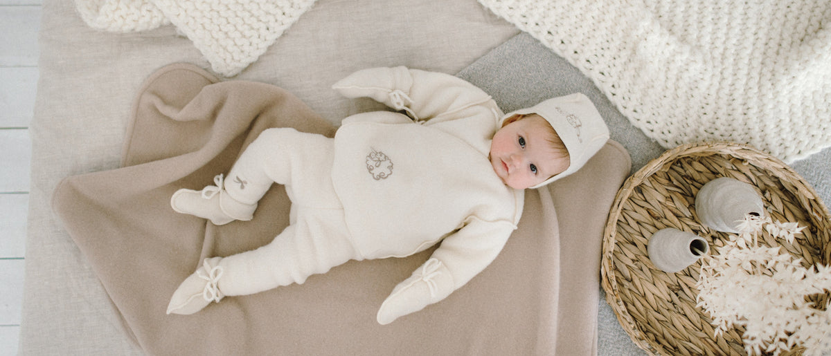 Baby in Merino-Strampler, Mütze und Schühchen auf einer weichen Decke